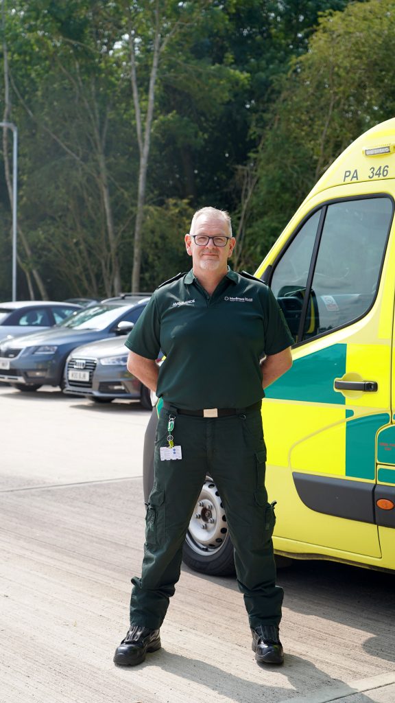 Singular ambulance staff posing in front of ambulance, he looks proud and is standing with his hands behind his back.