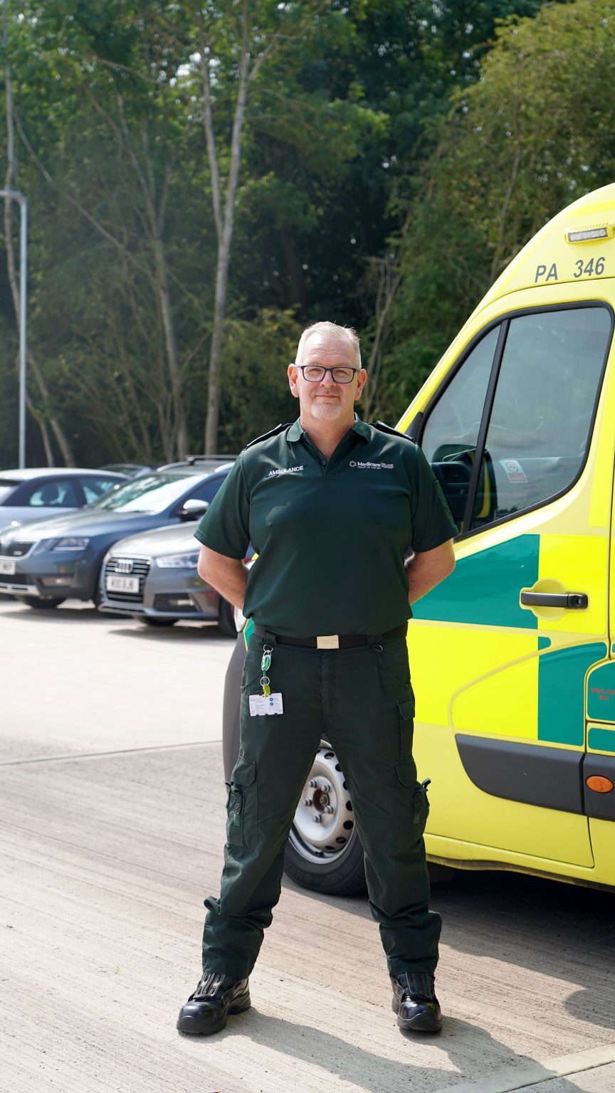 Singular ambulance staff posing in front of ambulance, he looks proud and is standing with his hands behind his back.