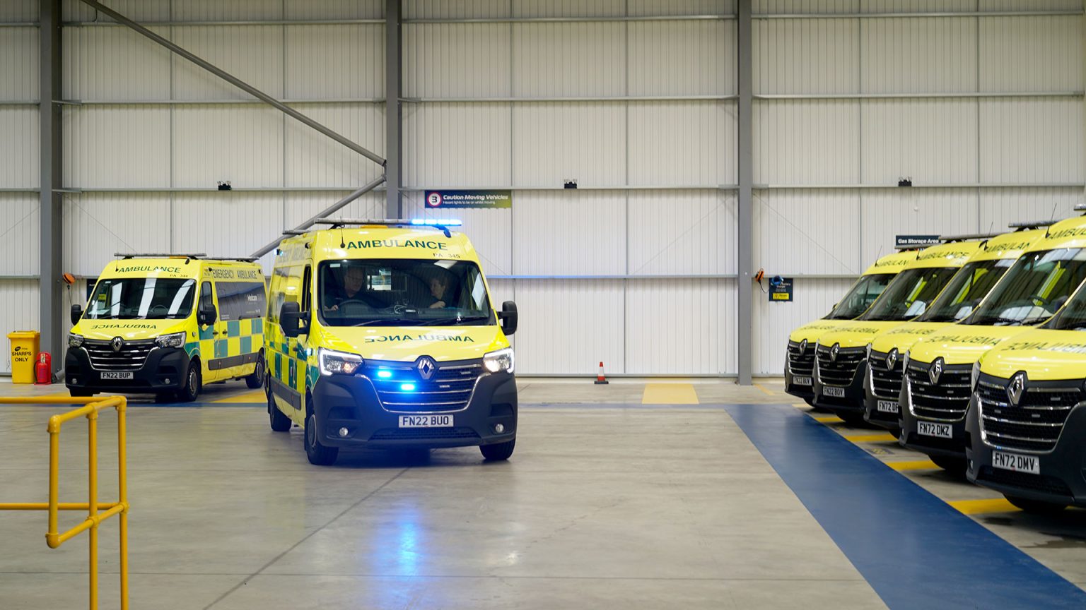 Ambulance parked up in what appears to be a warehouse of some kind. One ambulance at the front of the image has its blue flashing lights on. In this image there are two paramedics in the ambulance, they look ready to respond to an emergency. Different, further back angle.