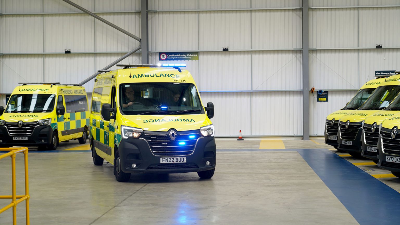 Ambulance parked up in what appears to be a warehouse of some kind. One ambulance at the front of the image has its blue flashing lights on. In this image there are two paramedics in the ambulance, they look ready to respond to an emergency.