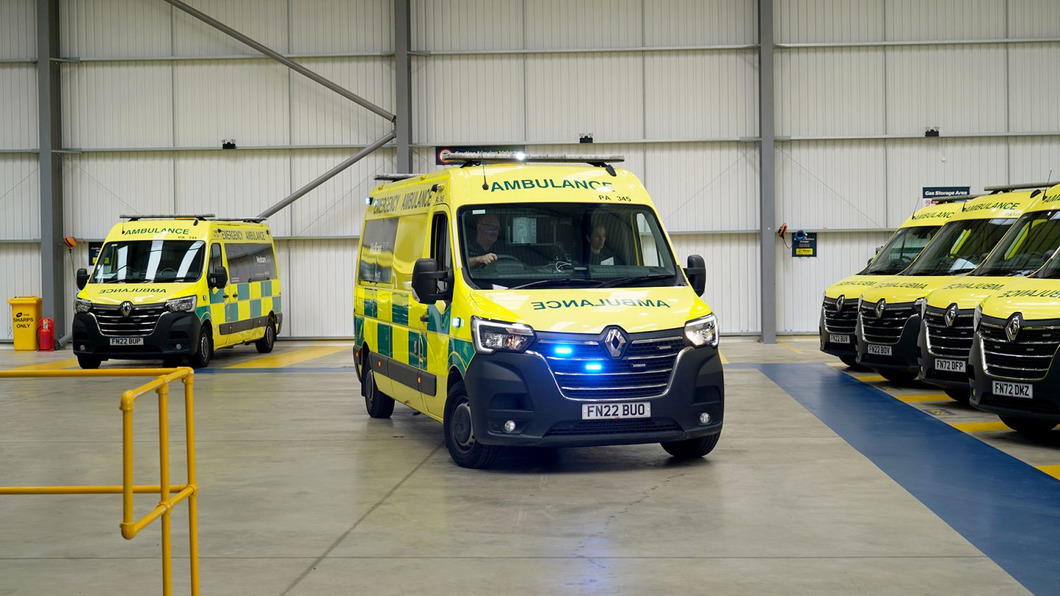 Ambulance parked up in what appears to be a warehouse of some kind. One ambulance at the front of the image has its blue flashing lights on. In this image there are two paramedics in the ambulance, they look ready to respond to an emergency.
