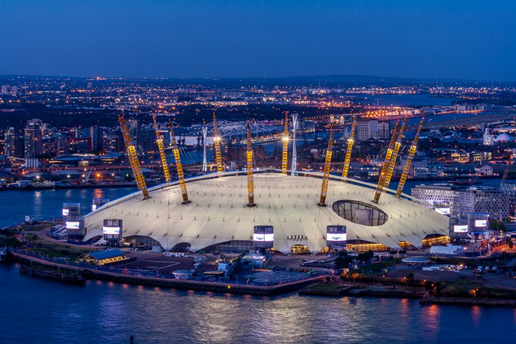 The O2 Arena at Night. The lights of the arena and the rest of London really light up, you can partially see the Thames, the water looks very calm and still.