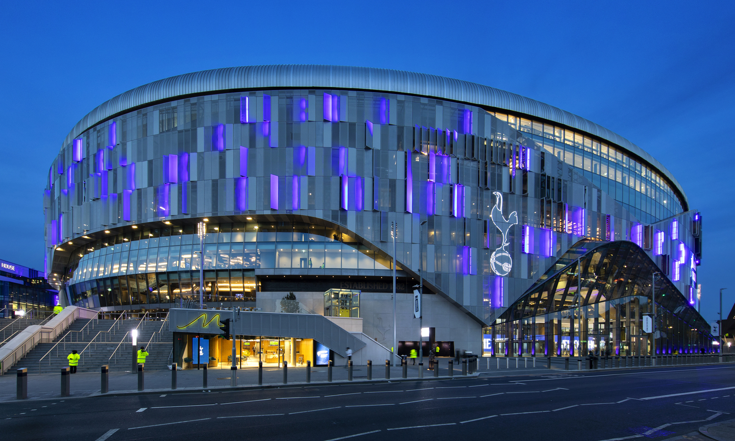 Tottenham Hotspur Football stadium at evening time. You can see some purple lights which look quite unique. There are many bollards along the road which runs alongside the stadium. There is also a large glass window area covering a side of the stadiums entrance.