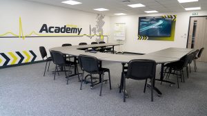 boardroom with large tv and medicare academy ems logo on wall. Some chairs and a table are in the centre